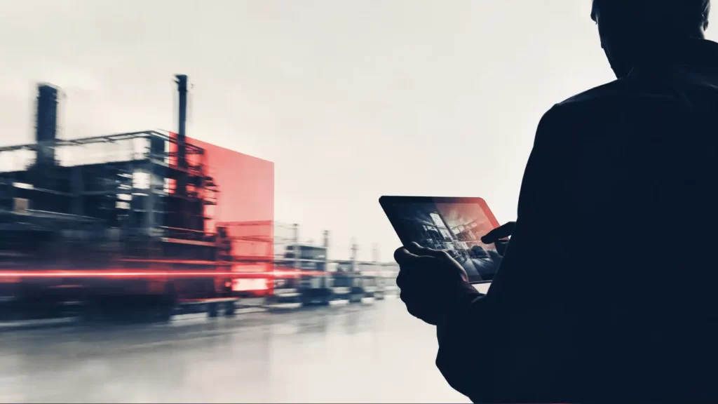 Uomo in controluce che guarda dati su un tablet davanti a uno stabilimento industriale, per rappresentare il marketing digitale per le aziende manifatturiere